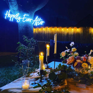 An outdoor evening setting with a 'Happily Ever After' neon sign glowing in blue, mounted on a tree trunk.