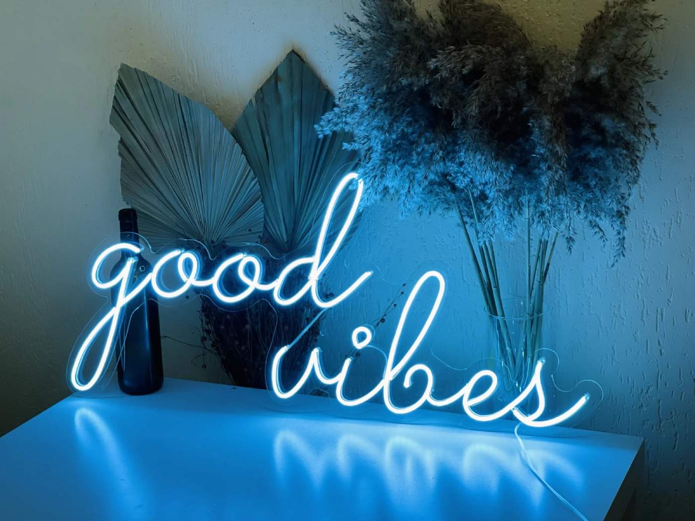 A neon sign with the phrase 'good vibes' in cursive, glowing in a bright blue color, positioned against a wall above a small white table.